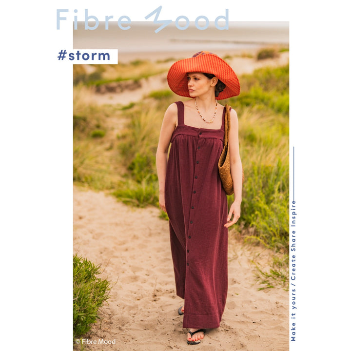 Woman in a long burgundy dress and red hat walking on a sandy path with grass, wearing a 'Fibre mood' branded outfit.