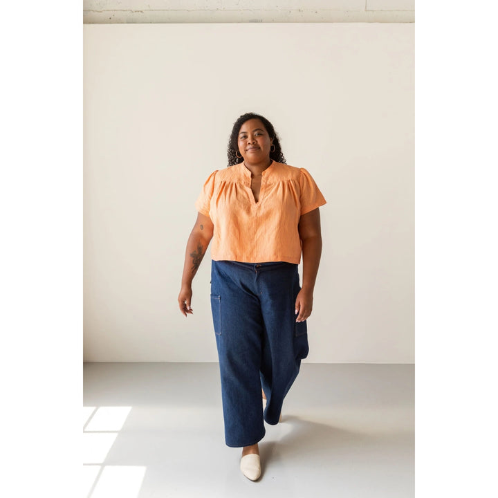 A woman modeling a short-sleeve peach-orange blouse with a gathered neckline and dark blue wide-leg pants, standing in a minimalist studio with a warm off-white background.