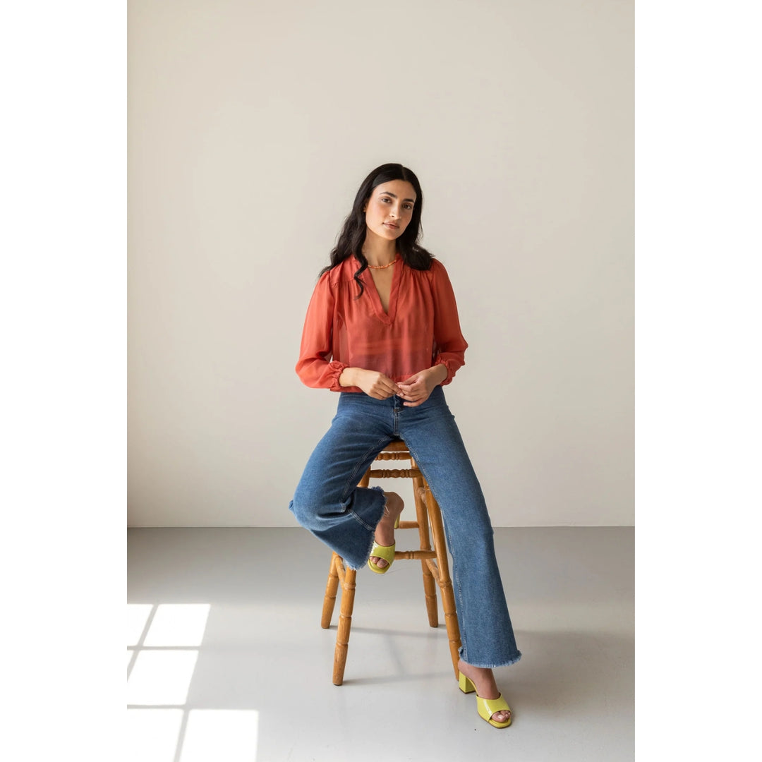 A woman sits on a wooden stool in a minimalist studio setting, wearing a rust-orange long-sleeve blouse, blue wide-leg jeans, and lime green sandals.