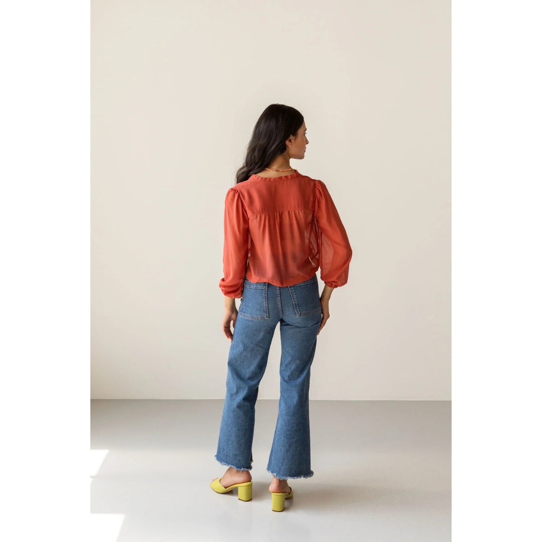 A woman is shown from behind wearing a rust-orange long-sleeve blouse, blue flare jeans, and yellow block-heel mules in a minimalist studio setting.