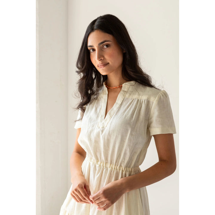 A woman wearing a short-sleeve cream-colored linen dress, standing in a brightly lit room with a white wall background.
