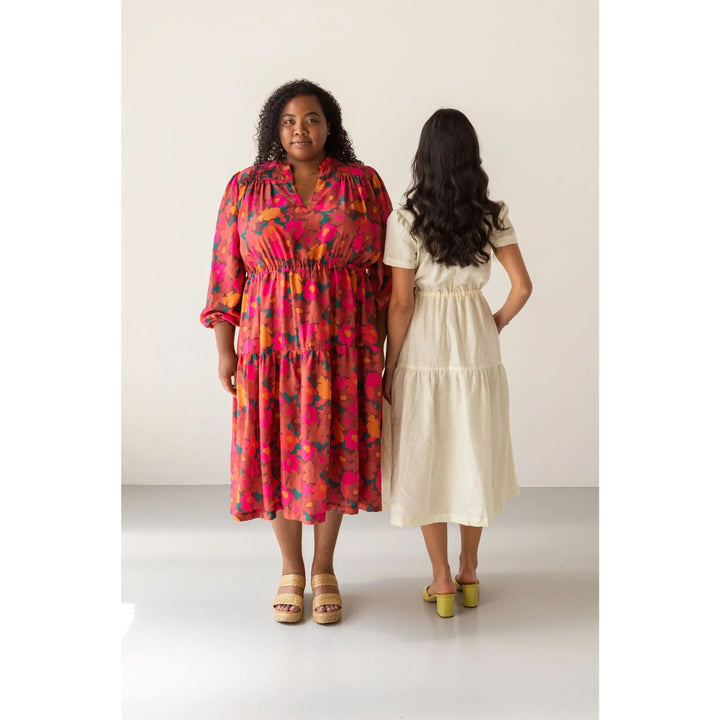 Two women modeling tiered midi dresses in a studio setting. The woman on the left faces forward wearing a vibrant pink, orange, and yellow abstract print dress with a smocked bodice and elbow-length sleeves.