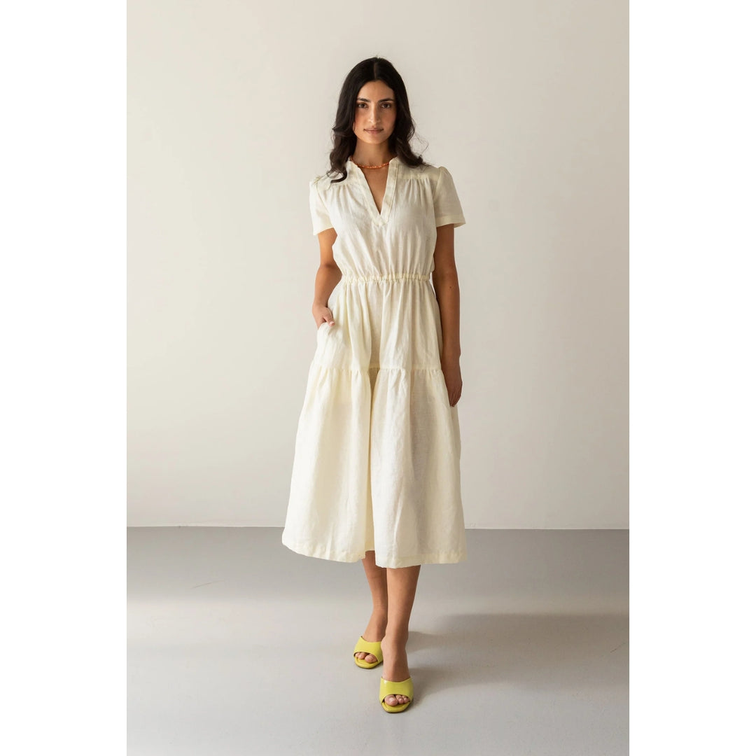 A woman modeling a short-sleeve midi dress in a warm off-white or cream color, paired with yellow slide sandals, standing against a neutral studio backdrop.