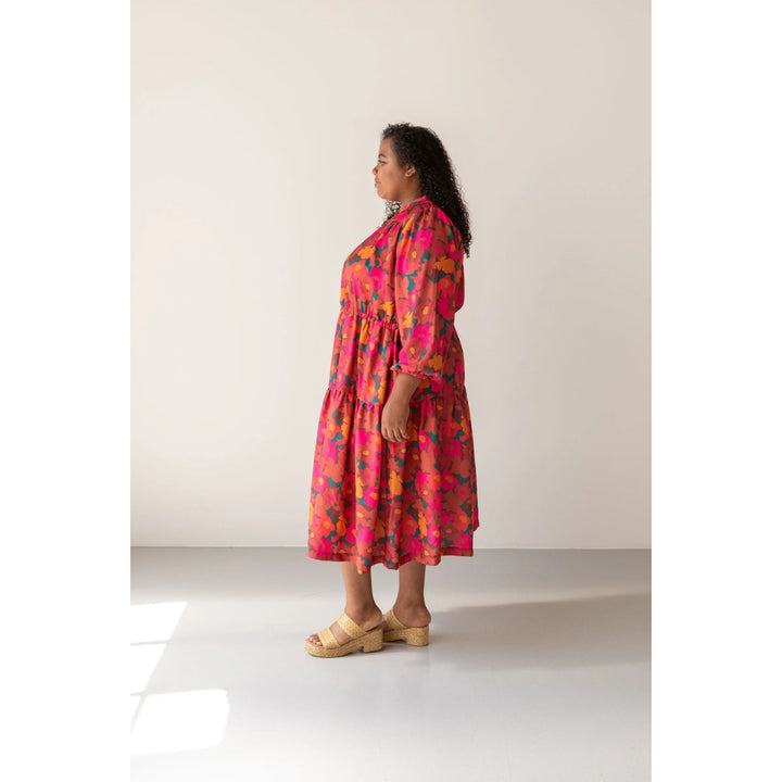 A woman stands in profile wearing a bright pink midi dress with a multicolor floral print in orange, yellow, and blue, paired with beige platform sandals, set against a minimalist cream studio background.