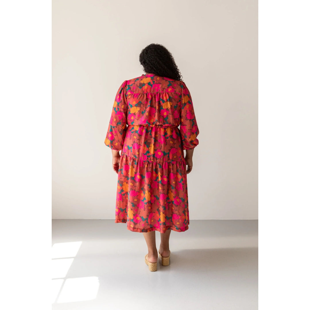A woman viewed from behind wearing a tiered midi dress with a vibrant pink, orange, and yellow floral pattern, standing in a minimalist studio setting.