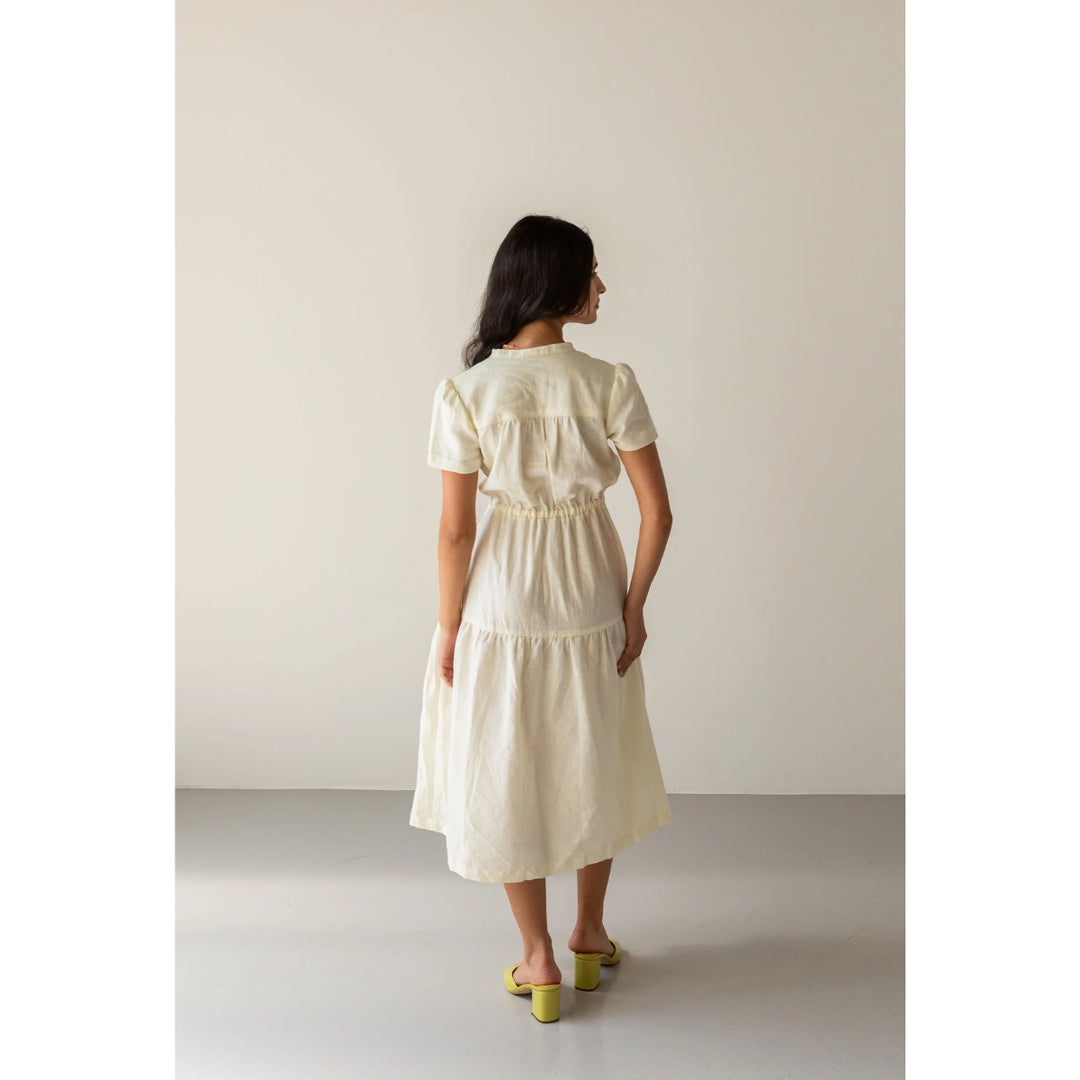 A woman viewed from behind wearing a short-sleeve cream-colored midi dress with a gathered waist, paired with lime green block-heel sandals, set against a minimalist beige studio background.