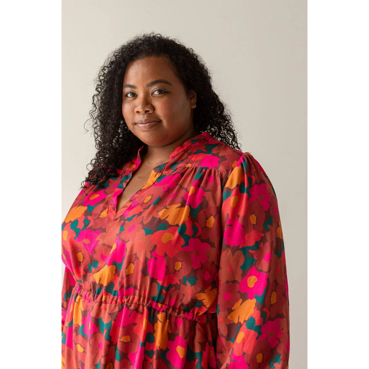 A woman with curly black hair wearing a long-sleeve, V-neck dress with a vibrant abstract floral pattern in pink, orange, and teal, set against a plain beige background.