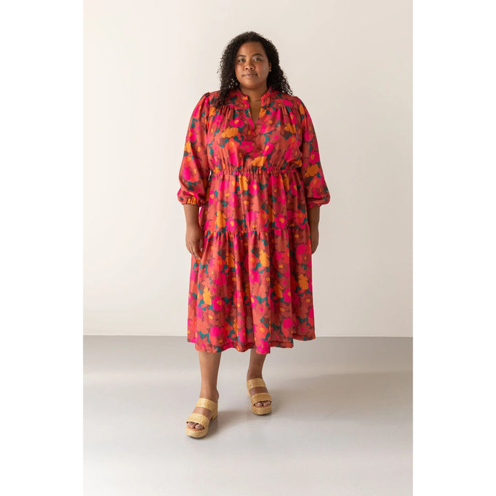 A woman modeling a tiered midi dress with a vibrant pink, orange, and yellow floral pattern, paired with beige platform sandals, set against a plain light beige studio background.
