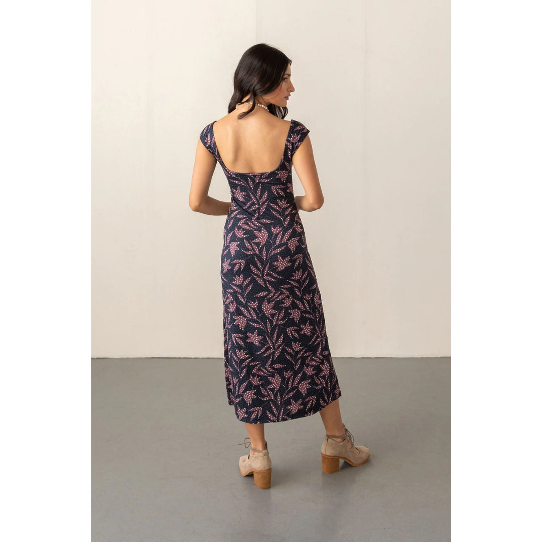 A woman stands with her back to the camera wearing a navy midi dress with a pink floral pattern and a square open back, paired with beige lace-up ankle boots, in front of a plain off-white wall.
