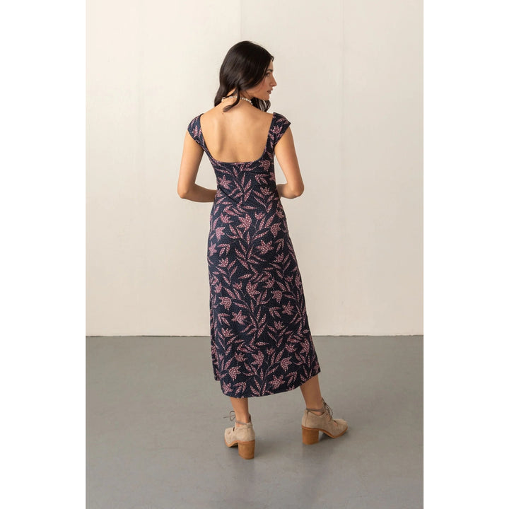 A woman stands with her back to the camera wearing a navy midi dress with a pink floral pattern and a square open back, paired with beige lace-up ankle boots, in front of a plain off-white wall.