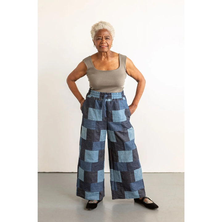 An older woman stands with hands on hips wearing wide-leg patchwork pants in shades of blue and navy, paired with a gray sleeveless top and black flats, against a plain white studio background.