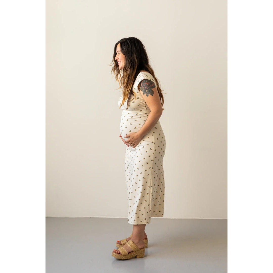 A pregnant woman stands in profile wearing a cream-colored short-sleeve maternity dress with a small brown polka dot pattern and tan wedge sandals, set against a minimalist beige studio background.