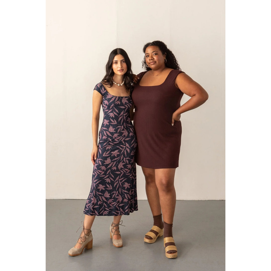 Two women modeling sleeveless dresses in a studio setting. The woman on the left wears a dark navy midi dress with a pink abstract leaf pattern, and the woman on the right wears a solid dark brown scoop-neck mini dress.