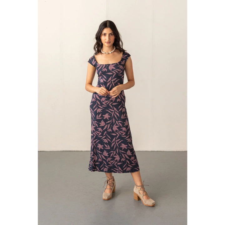 A woman modeling a navy blue midi dress with a pink floral pattern, styled with beige lace-up heels and a gold necklace, standing against a plain off-white wall and gray floor.