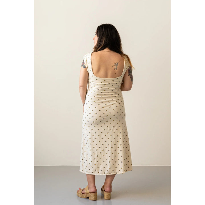 A woman viewed from behind wearing a cream-colored midi dress with a small brown floral pattern, standing against a plain beige studio background.