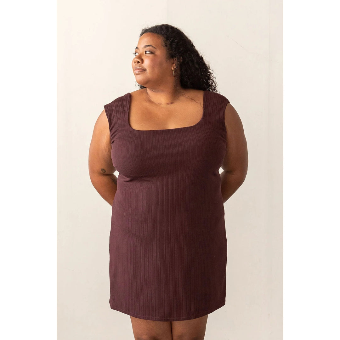 A woman modeling a sleeveless, scoop-neck ribbed dress in a deep plum-brown color against a plain off-white background.