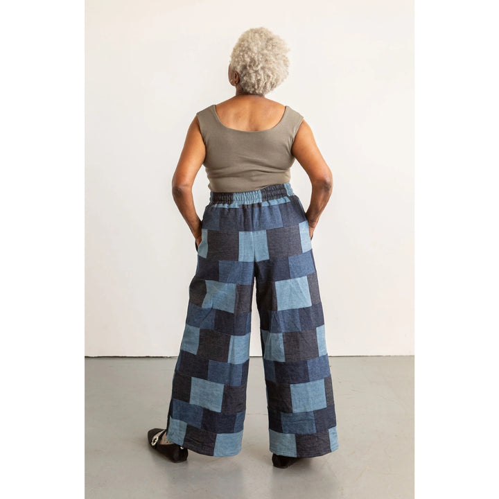An older woman viewed from behind wearing a sleeveless taupe top and wide-leg patchwork pants in shades of blue, navy, and gray, standing against a plain off-white wall.