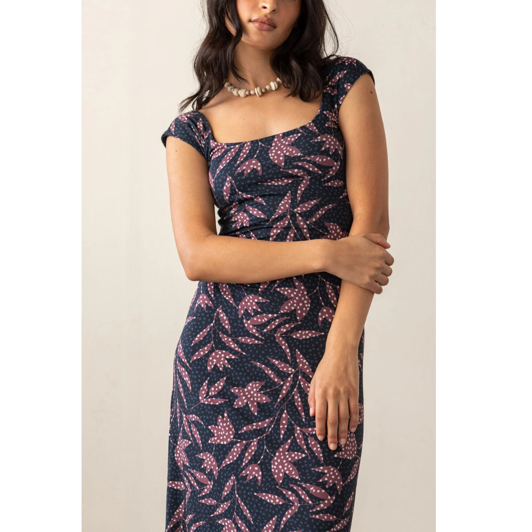 A woman wearing a navy midi dress with a pink and white leaf pattern, styled with a chunky pearl necklace against a plain beige background.