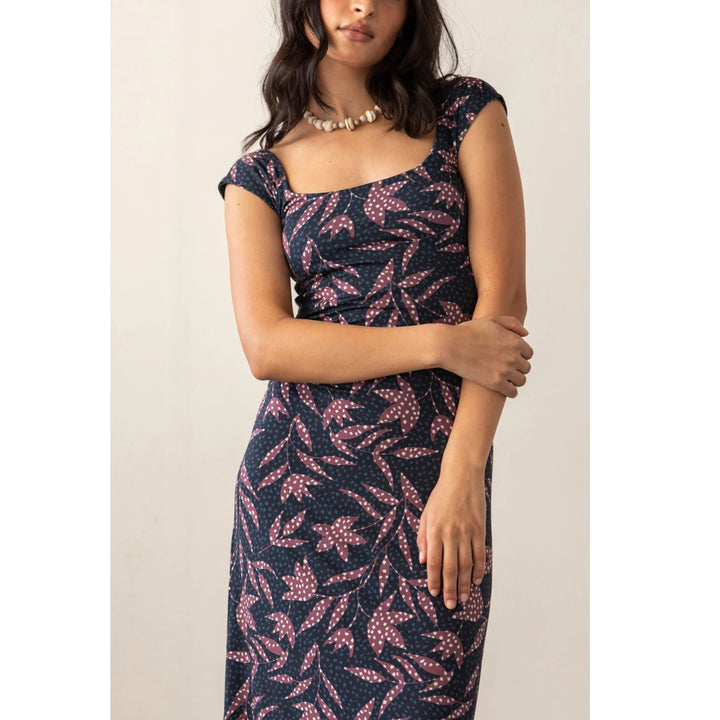A woman wearing a navy midi dress with a pink and white leaf pattern, styled with a chunky pearl necklace against a plain beige background.