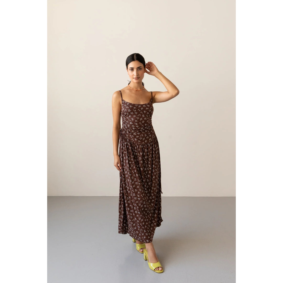 A woman modeling a brown spaghetti-strap maxi dress with a small white floral pattern, paired with yellow high-heeled sandals, set against a minimalist beige and gray studio background.