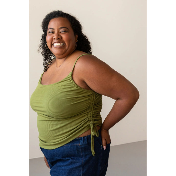 A woman smiling and posing in a ribbed olive green tank top with side ruching and tie details, paired with blue jeans, against a plain beige studio background.