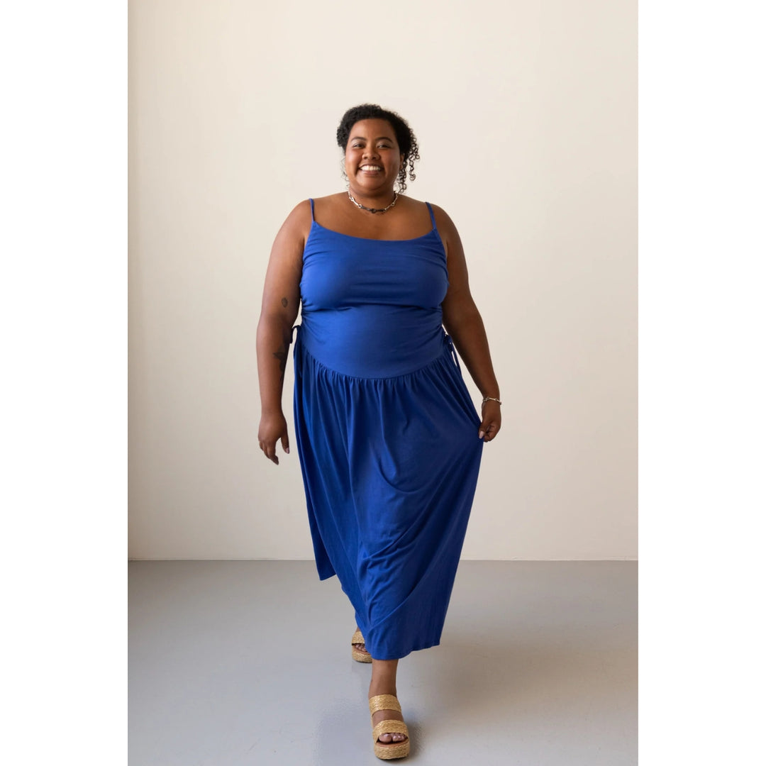 A woman modeling a bright royal blue sleeveless midi dress with thin straps and a gathered skirt, standing against a plain light beige studio background.