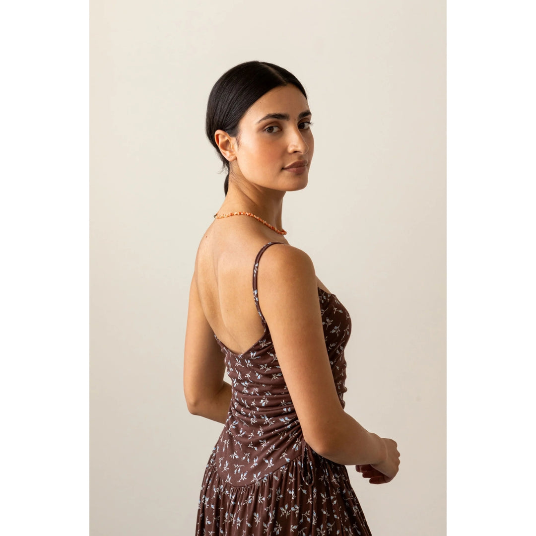 A woman wearing a brown spaghetti-strap dress with a small white floral pattern, photographed from the back and side against a plain beige studio background.