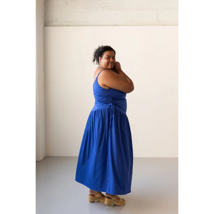 A woman modeling a bright blue sleeveless maxi dress with a ruched bodice and side tie, paired with tan wedge sandals, standing in a minimalist studio with a cream-colored wall.