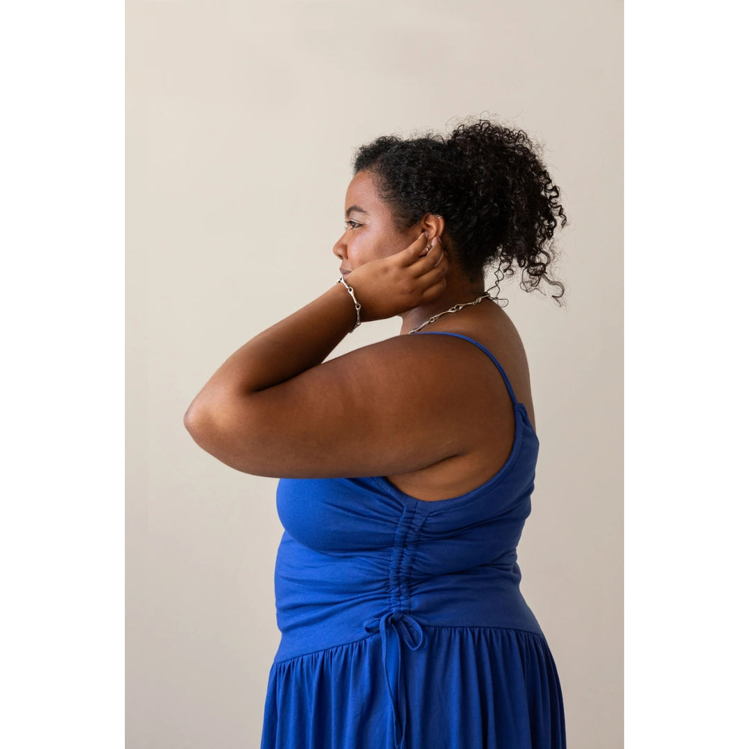 A woman in profile wearing a bright blue sleeveless dress with a gathered back and side tie, photographed against a plain light beige studio background.