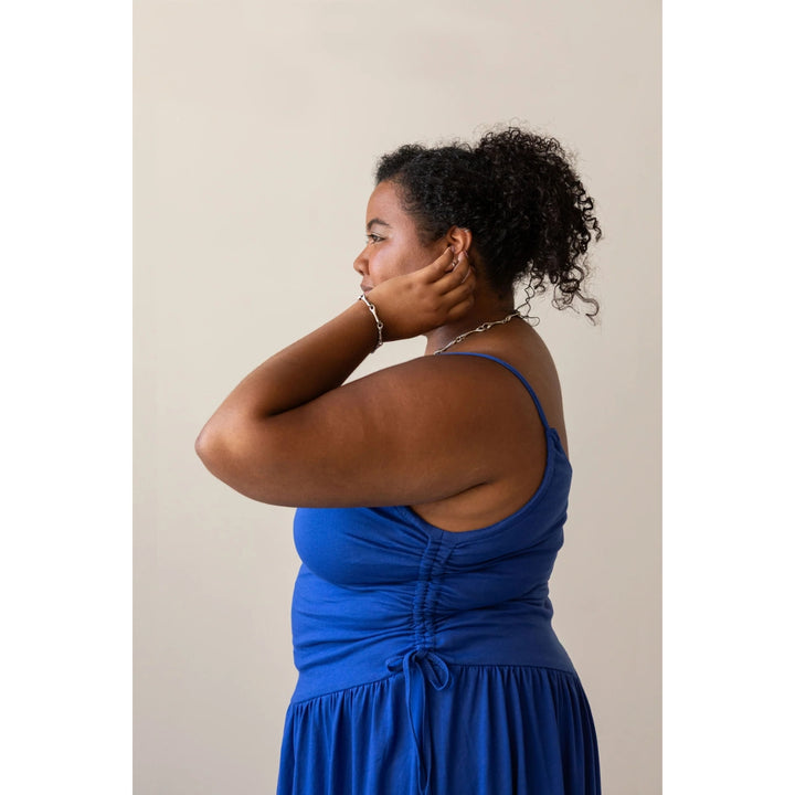 A woman in profile wearing a bright blue sleeveless dress with a gathered back and side tie, photographed against a plain light beige studio background.