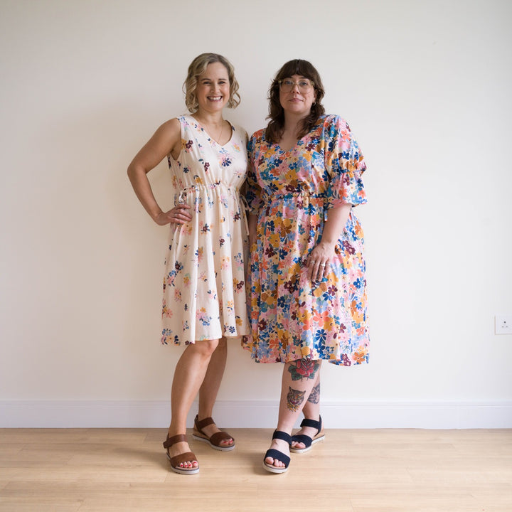 Two women wearing floral dresses standing against a plain wall.