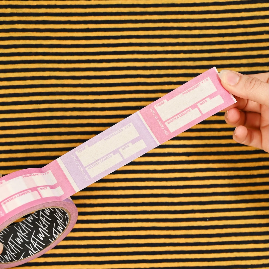 Pink and lavender washi tape with white text and boxes, shown partially unrolled from a roll with a hand holding the end, set against a black and yellow striped background.