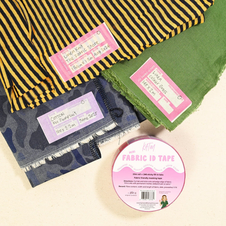 A roll of pink 'Kylie and the machine' Fabric ID Tape is displayed on a light beige surface, surrounded by folded fabric samples labeled with matching pink tags. The fabrics include a yellow and black striped linen, a green cotton, a blue camouflage cotton, and a dark gray frayed cotton.