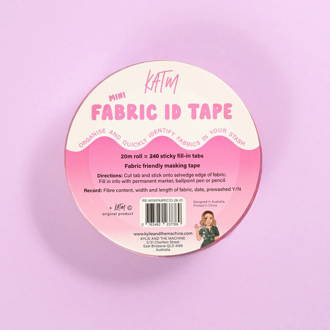 A round spool of KATM Mini Fabric ID Tape, featuring a pink and cream label with instructions and a barcode, set against a solid pastel pink background.