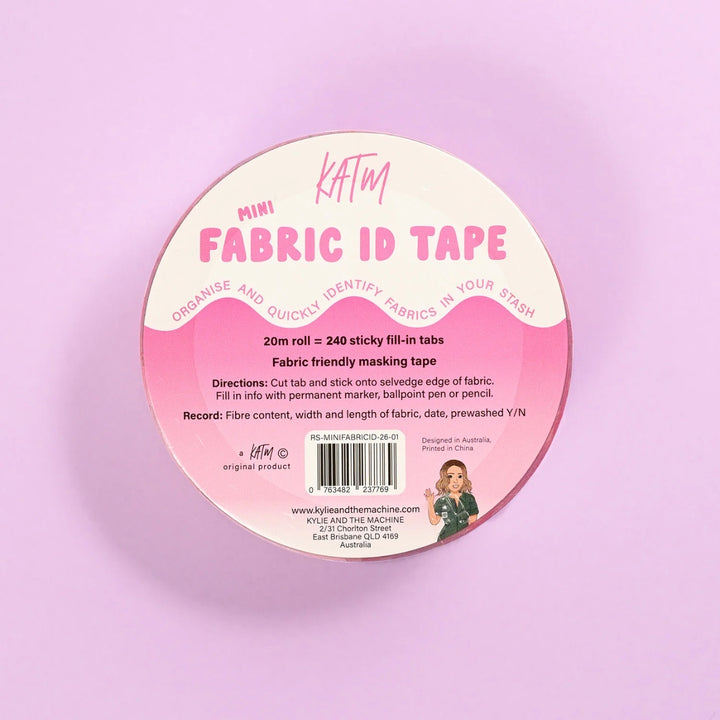 A round spool of KATM Mini Fabric ID Tape, featuring a pink and cream label with instructions and a barcode, set against a solid pastel pink background.