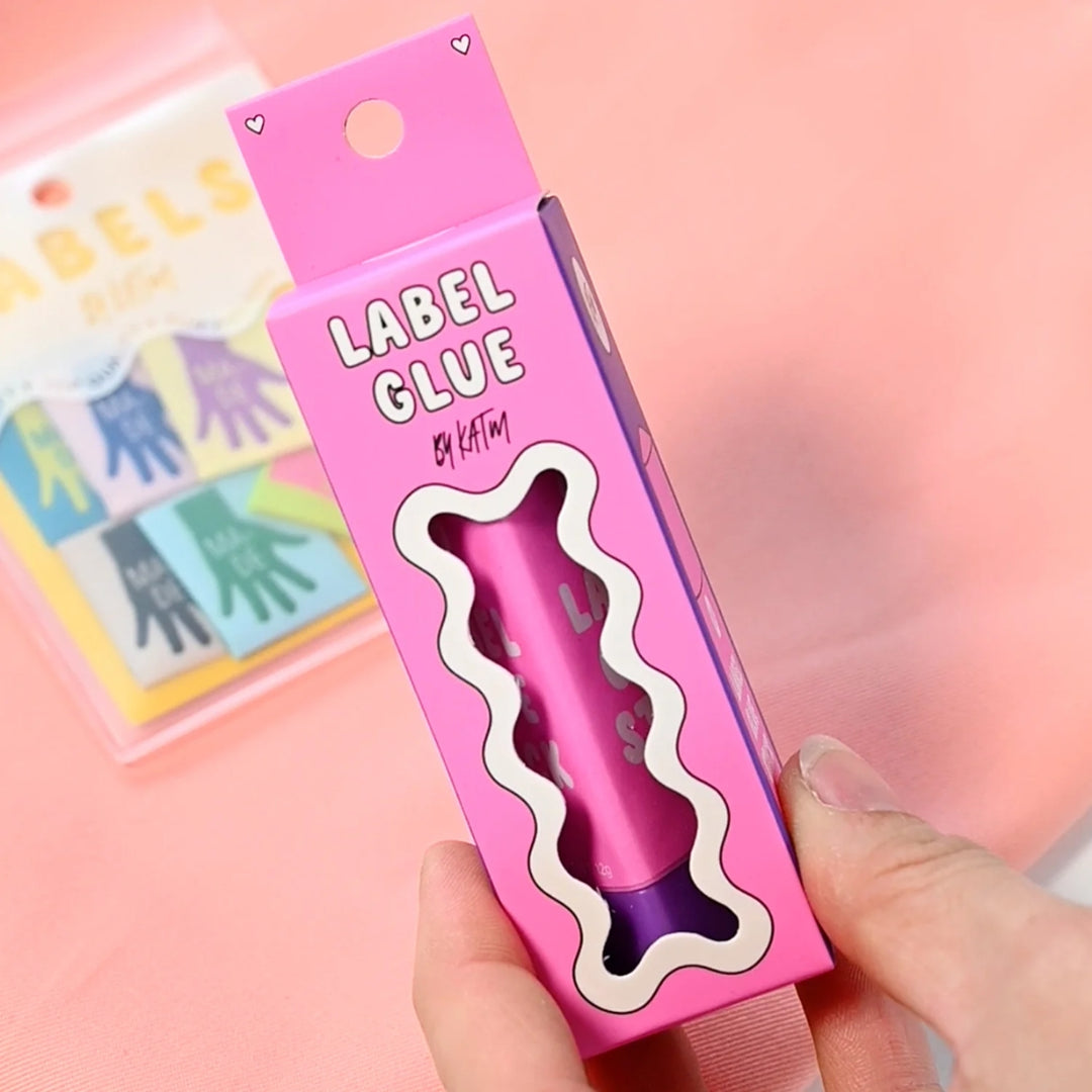 A hand holding a pink box of 'LABEL GLUE' by Kylie and the Machine, showing the purple glue stick through a wavy cutout window, with a pink background and a blurred card in the background.