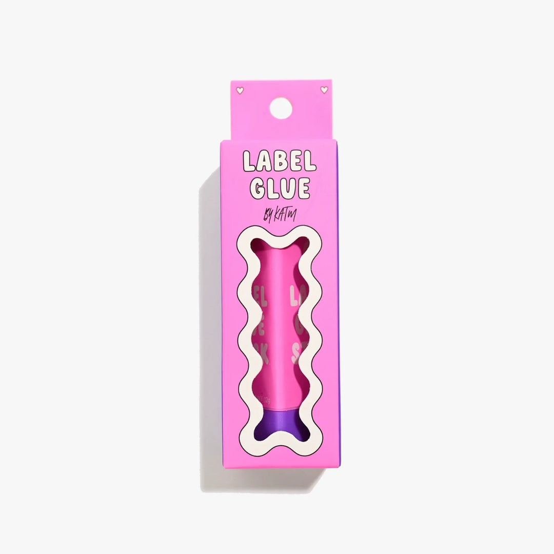 A pink bone-shaped label glue stick in its retail packaging, featuring a wavy cutout window that reveals the pink and purple product inside. The packaging is bright pink with the text 'LABEL GLUE' and 'by kylie and the machine' visible.