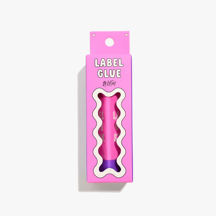 A pink bone-shaped label glue stick in its retail packaging, featuring a wavy cutout window that reveals the pink and purple product inside. The packaging is bright pink with the text 'LABEL GLUE' and 'by kylie and the machine' visible.