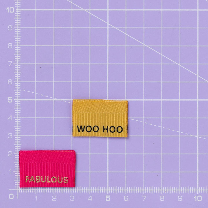 Two woven labels, one yellow with 'WOO HOO' and one pink with 'FABULOUS', on a purple grid background.
