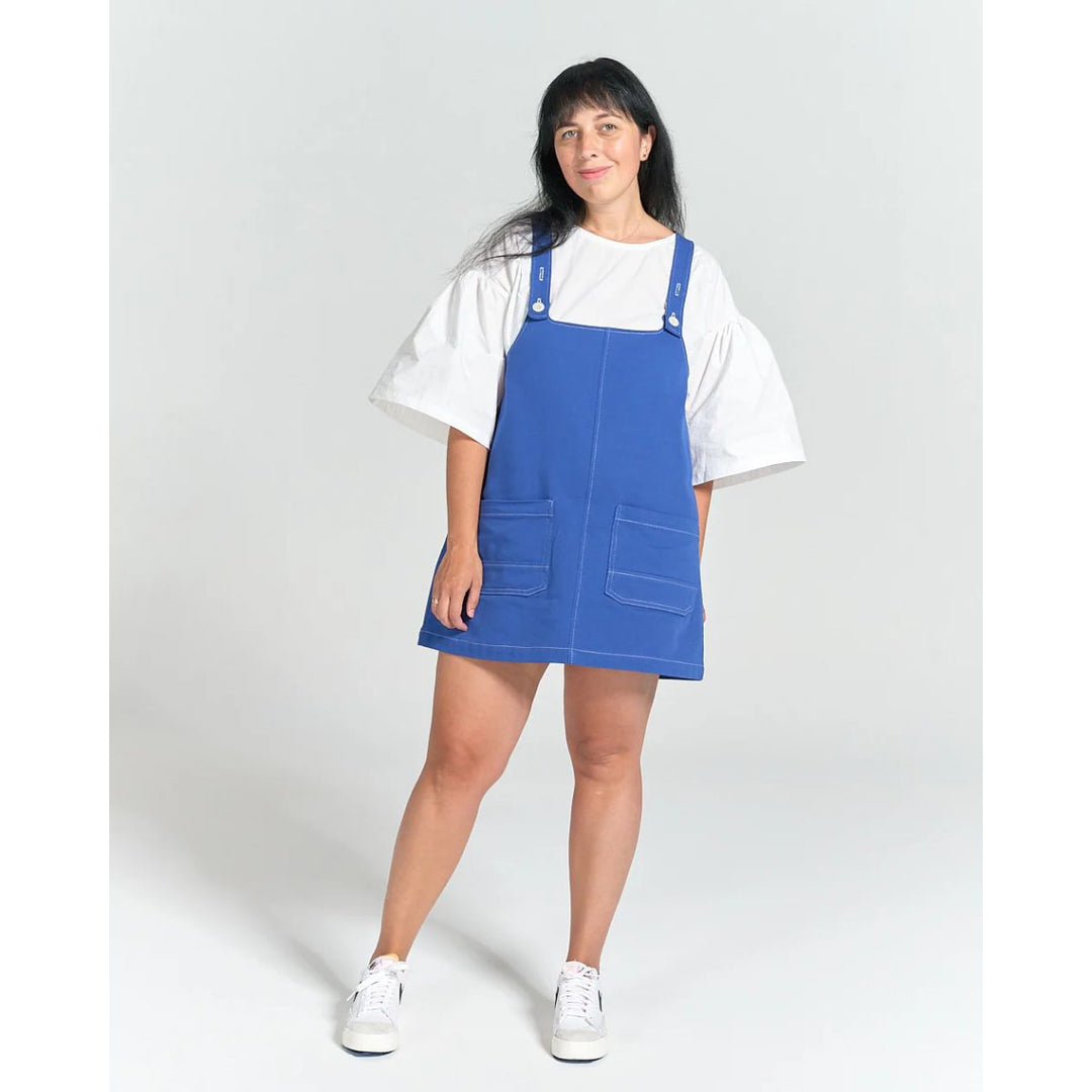 Person wearing a blue pinafore dress over a white shirt on a light gray background