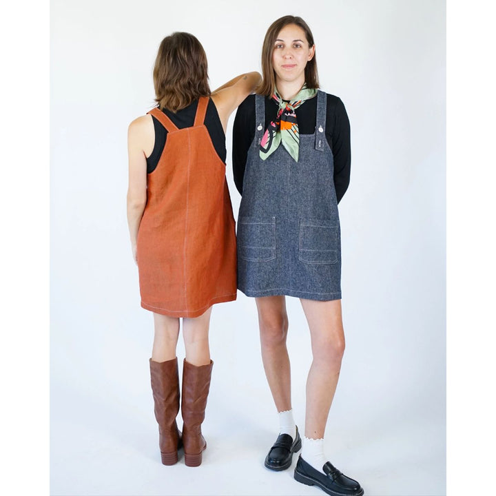 Two women wearing pinafore dresses, one orange and one blue, standing back-to-back against a white background.