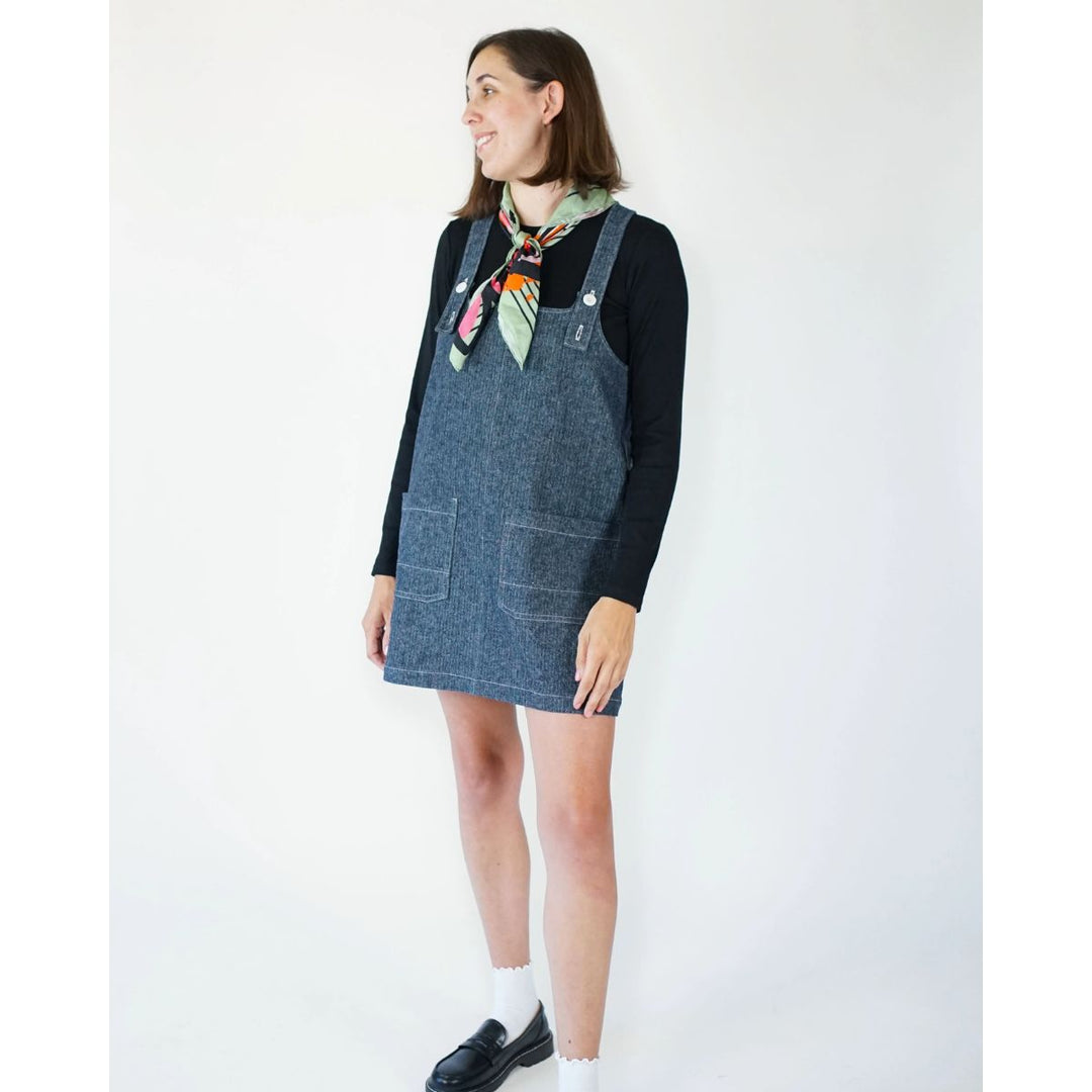 Woman wearing a denim pinafore dress over a black long-sleeve shirt with a colorful scarf on a white background