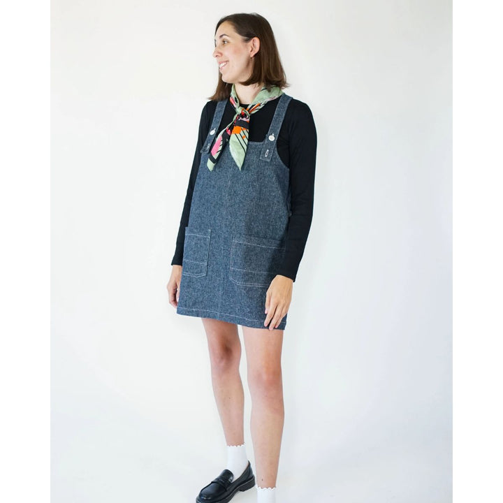 Woman wearing a denim pinafore dress over a black long-sleeve shirt with a colorful scarf on a white background