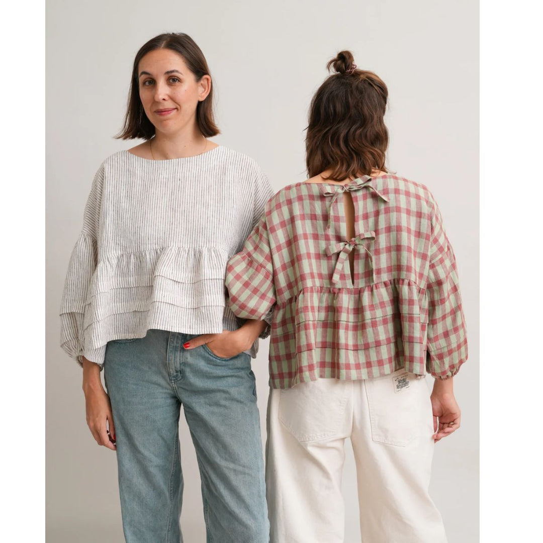 Two women wearing tops and pants, one facing the camera and the other back to the camera, on a plain background.