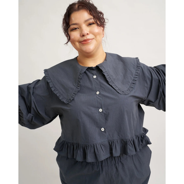 Woman wearing a dark blue blouse with ruffled collar and cuffs on a plain background