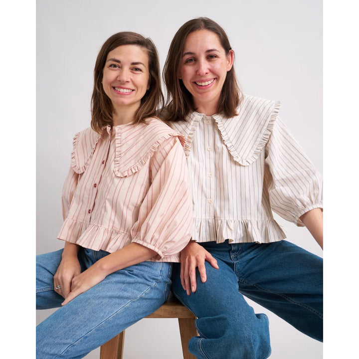 Two women wearing matching ruffled blouse and jeans sitting on a stool against a white background.