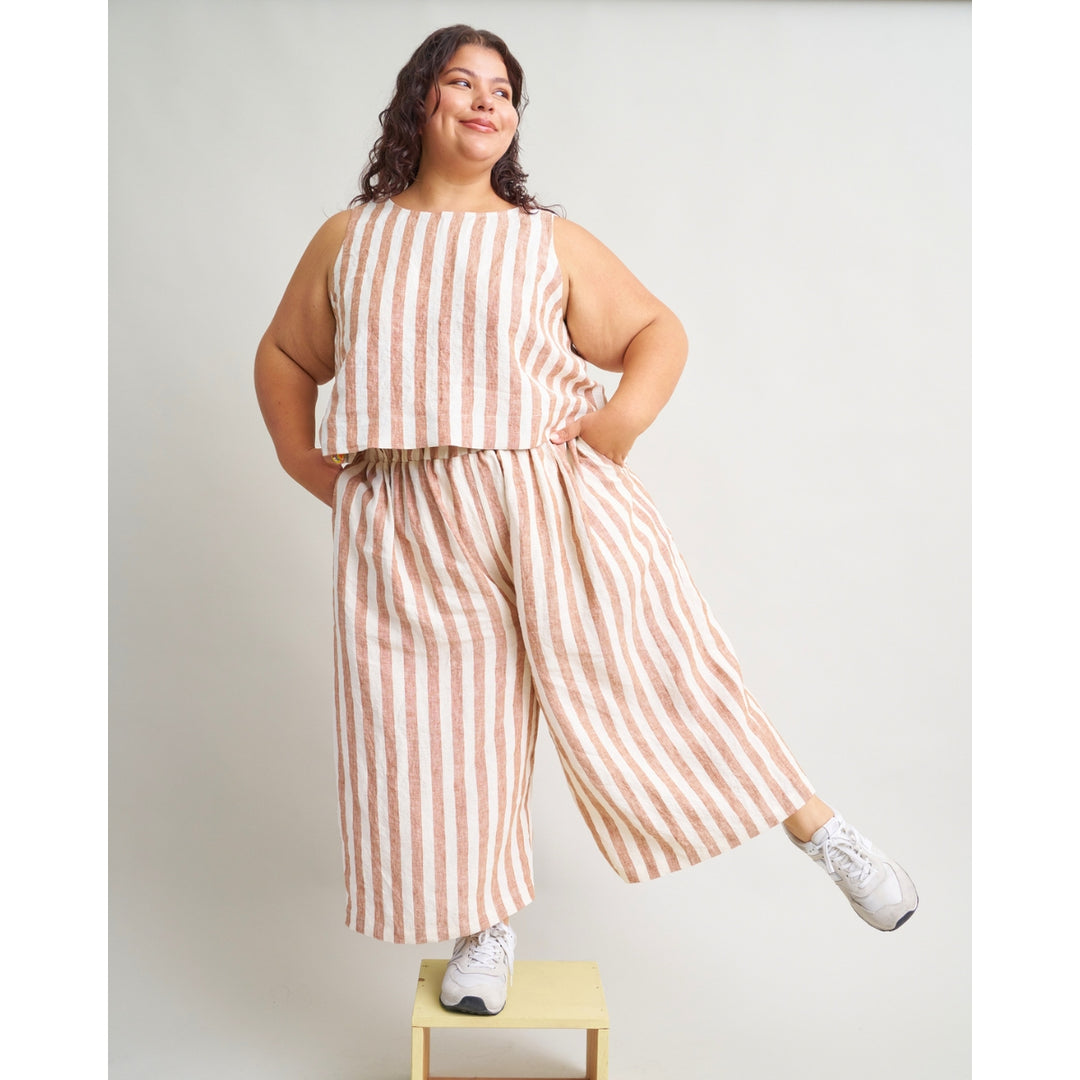 A woman modeling a sleeveless striped two-piece outfit with cropped top and wide-leg pants, standing on a small wooden stool against a light gray studio background.