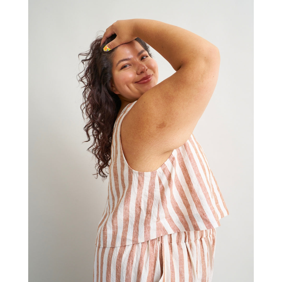 A woman with long dark curly hair poses in a sleeveless, tiered top with vertical pink and off-white stripes, set against a plain light gray background.