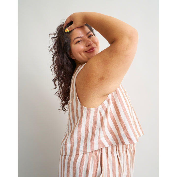 A woman with long dark curly hair poses in a sleeveless, tiered top with vertical pink and off-white stripes, set against a plain light gray background.