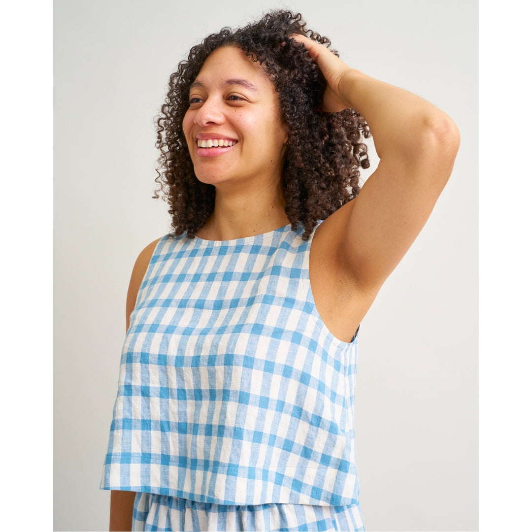 A woman with curly brown hair smiling and looking to the side, wearing a sleeveless blue and white gingham check top against a plain light gray background.